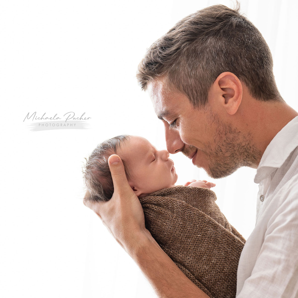 Vater hält sein neugeborenes Baby auf Augenhöhe und berührt liebevoll mit der Nase die Stirn des schlafenden Kindes.